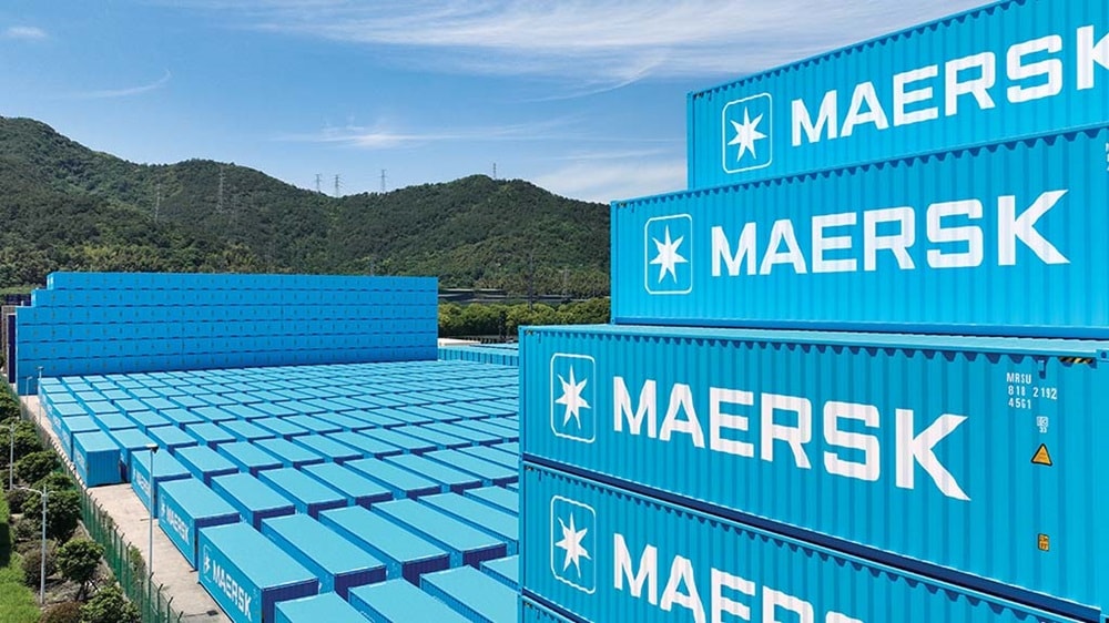 Stacked Maersk shipping containers in a large container yard with green hills in the background, representing global logistics and supply chain operations.