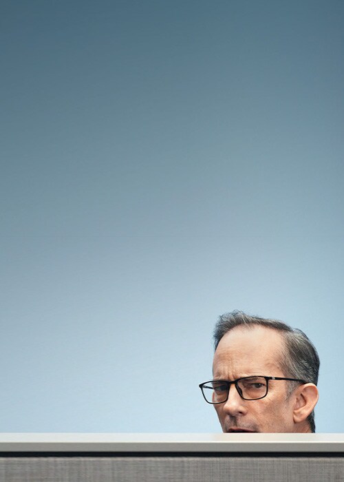 A man wearing glasses looks out from behind a cubicle wall, representing curiosity and teamwork in a business setting